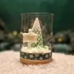 Ręcznie robiona świeca sojowa w kształcie zielonej choinki i tabliczki z napisem ‘Merry Christmas’, umieszczona w szklanym słoiku z drewnianą pokrywką i ozdobną podstawą