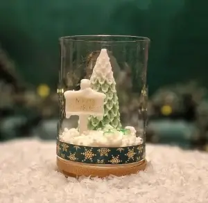Ręcznie robiona świeca sojowa w kształcie zielonej choinki i tabliczki z napisem ‘Merry Christmas’, umieszczona w szklanym słoiku z drewnianą pokrywką i ozdobną podstawą