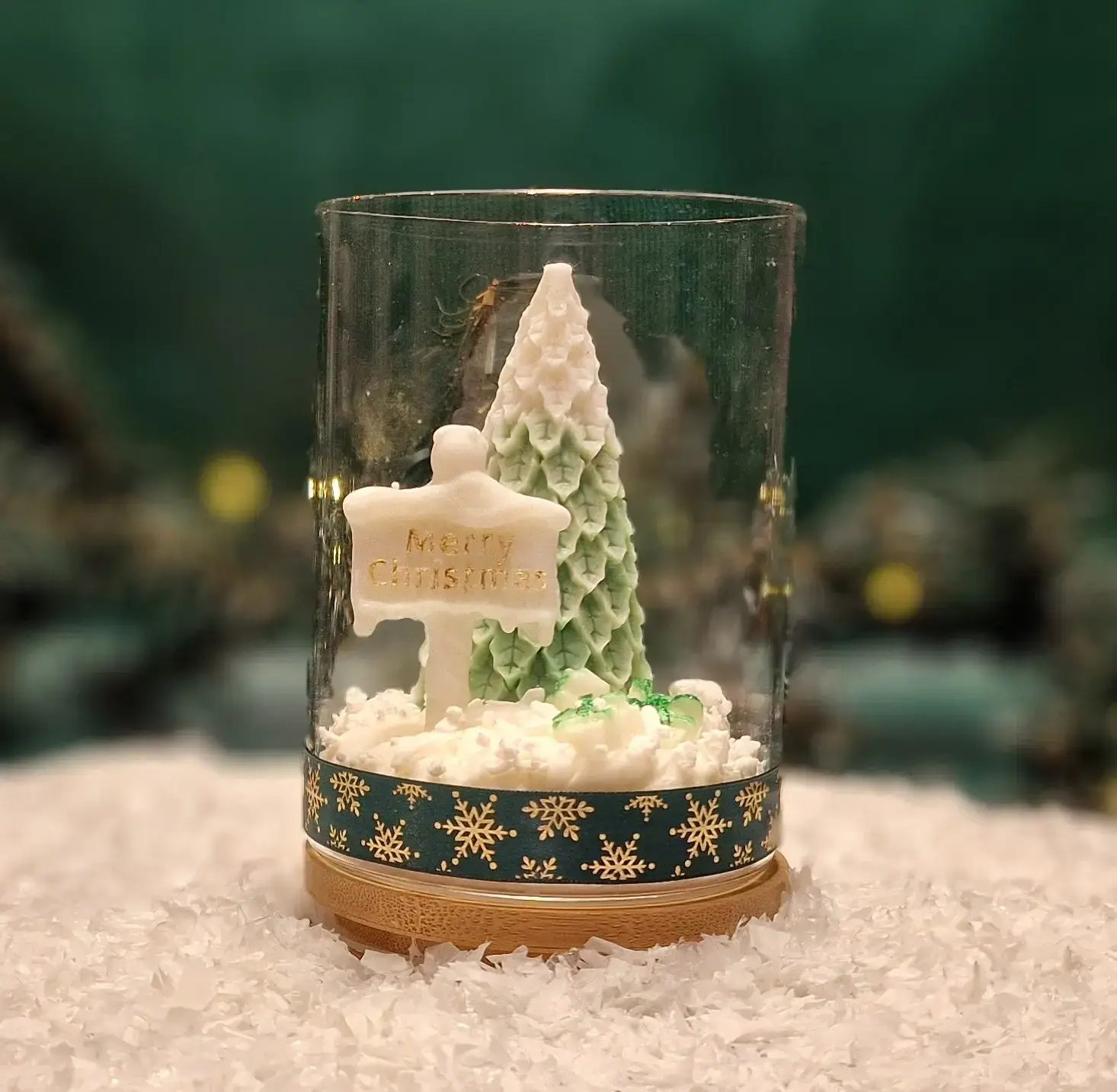 Ręcznie robiona świeca sojowa w kształcie zielonej choinki i tabliczki z napisem ‘Merry Christmas’, umieszczona w szklanym słoiku z drewnianą pokrywką i ozdobną podstawą