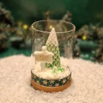 Ręcznie robiona świeca sojowa w kształcie zielonej choinki i tabliczki z napisem ‘Merry Christmas’, umieszczona w szklanym słoiku z drewnianą pokrywką i ozdobną podstawą