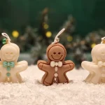 Ręcznie robione świece sojowe w kształcie ludków piernikowych: biały z miętowymi ozdobami, brązowy klasyczny oraz biały z białymi detalami, ustawione na sztucznym śniegu.