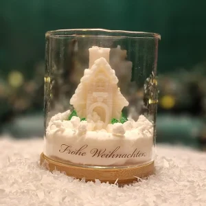 Handgemachte Sojawachskerze in Form eines verschneiten Häuschens mit Tannen, präsentiert in einem Glas mit Holzsockel und dem Schriftzug ‘Frohe Weihnachten’