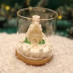 Handmade soy wax candle shaped like a snowy cottage with trees, displayed in a glass jar with a wooden base and the inscription ‘Frohe Weihnachten’.