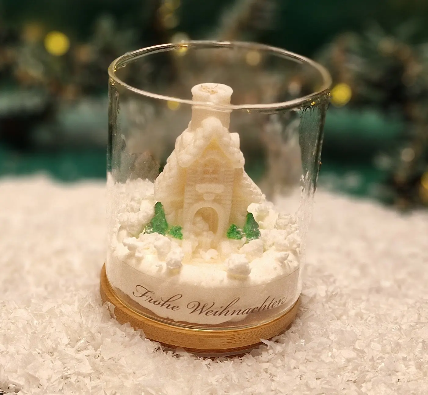 Handmade soy wax candle shaped like a snowy cottage with trees, displayed in a glass jar with a wooden base and the inscription ‘Frohe Weihnachten’. Handmade soy wax candle shaped like a snowy cottage with trees, displayed in a glass jar with a wooden base and the inscription ‘Frohe Weihnachten’.