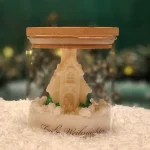 Handmade soy wax candle shaped like a winter cottage, placed in a glass jar with a wooden lid and the inscription ‘Frohe Weihnachten’.